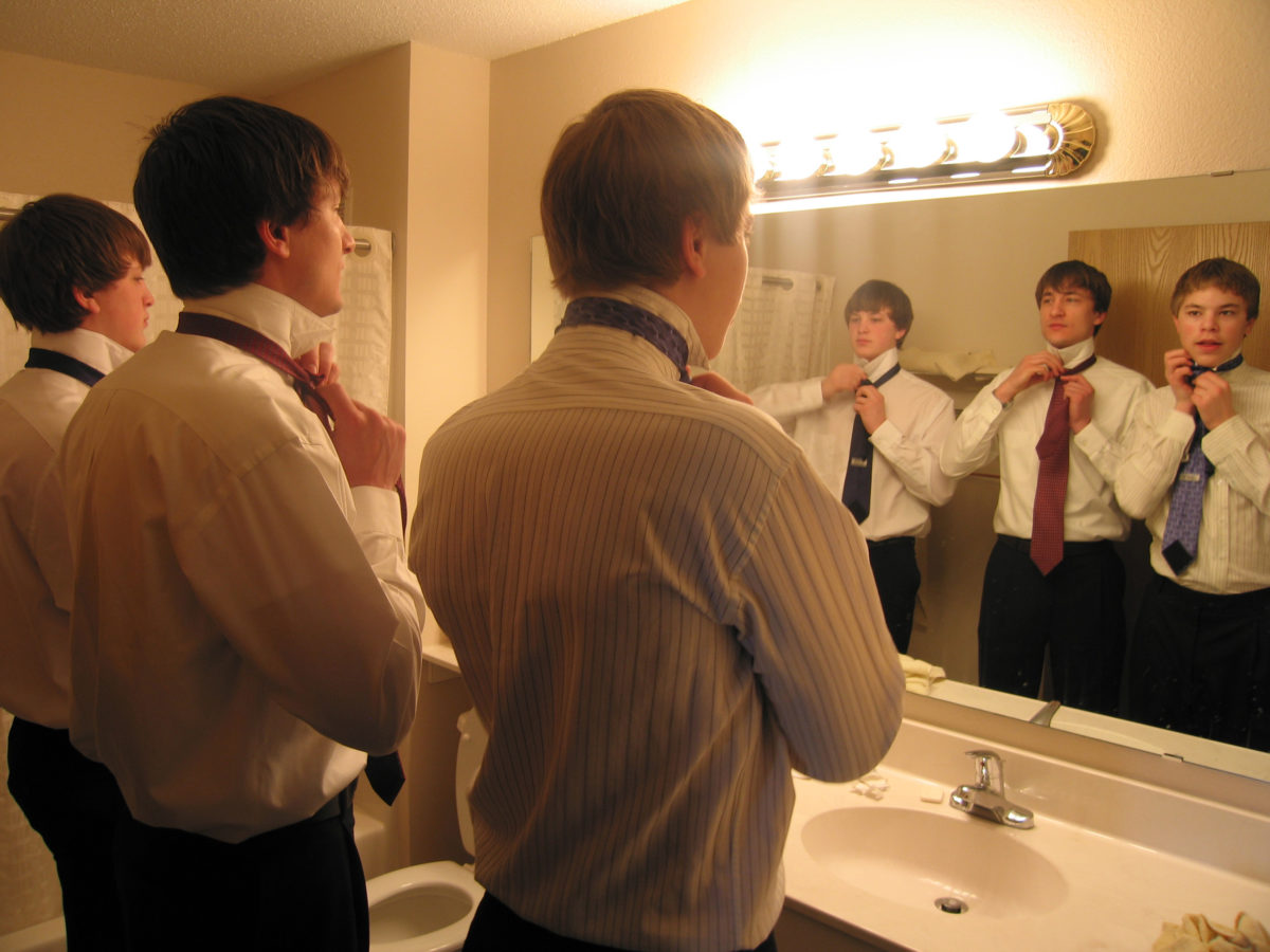 Ben showing Joe and Rob how to tie ties, reflected in a mirror