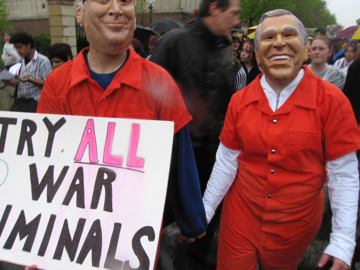 Protestors in Bush and Cheney masks, in orange prison jumpsuits. Sig says, "Try All War Criminals."