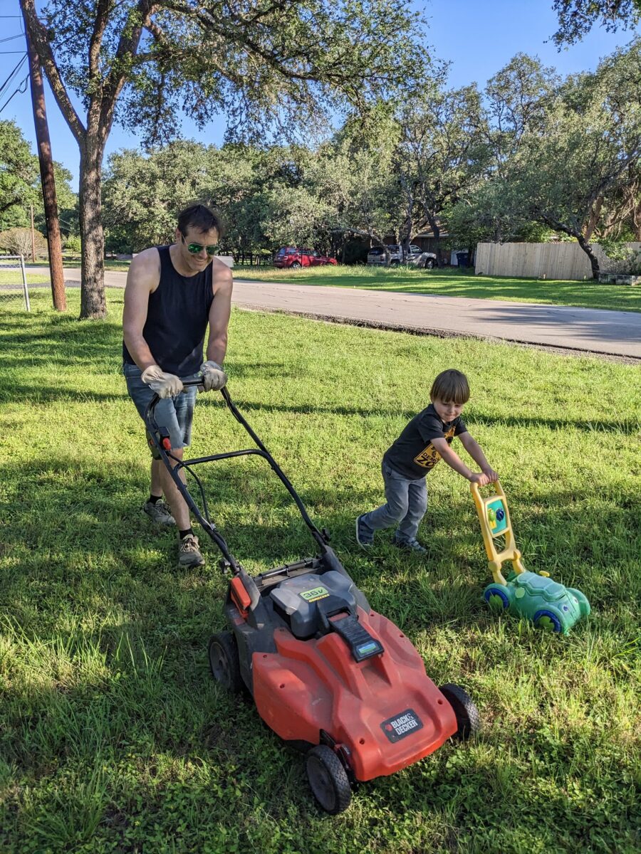 Man mows the lawn with a boy with toy lawnmower beside him.