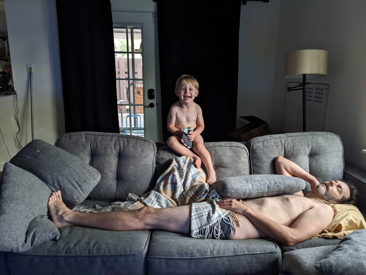 A man sleeps on a couch with a young boy laughing.