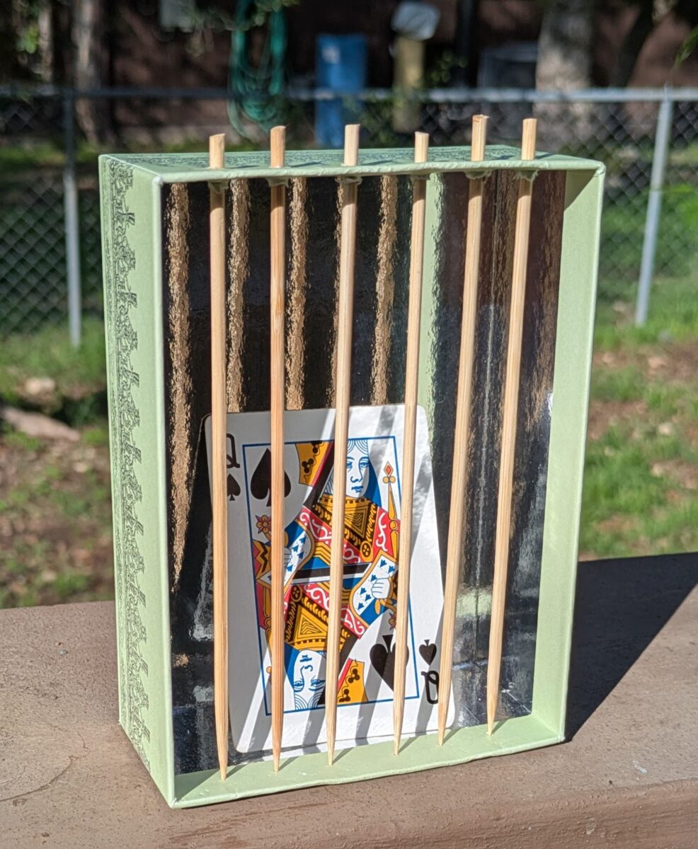 The queen of spades is trapped in a cardboard jail with wooden skewer prison bars.